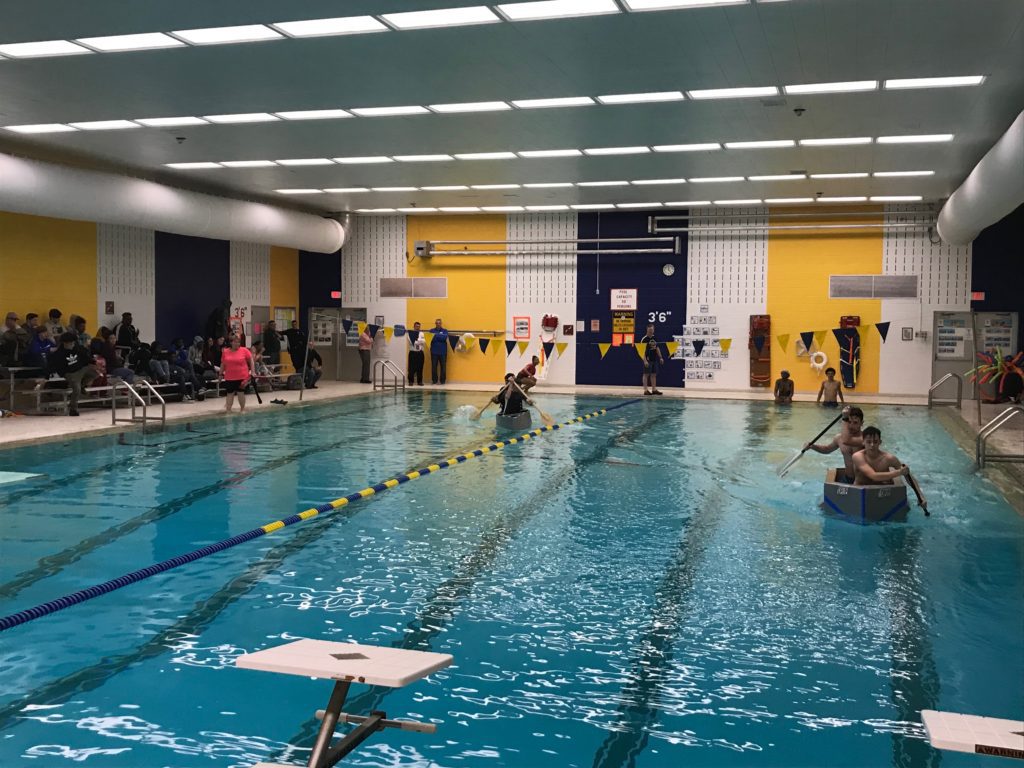 students paddle in cardboard boats across indoor swimming pool