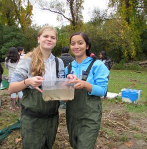 two girls in water waders holding a container with river water