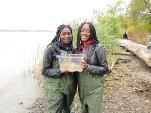 two girls in water waders holding a container with river water