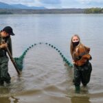 two high school students wearing chest waders walk a seine net out of the river