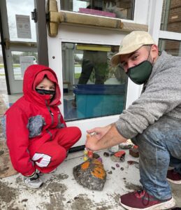 a man and a young boy on the school sidewalk painting a garden rock