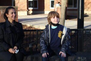 Dr. Suttmeier sits next to an elementary boy on a bench outisde the school