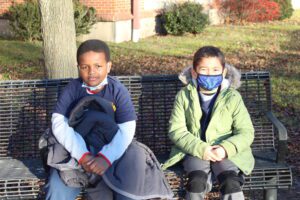 an african american boy and hispanic boy sit next to each other on a bench outside the school