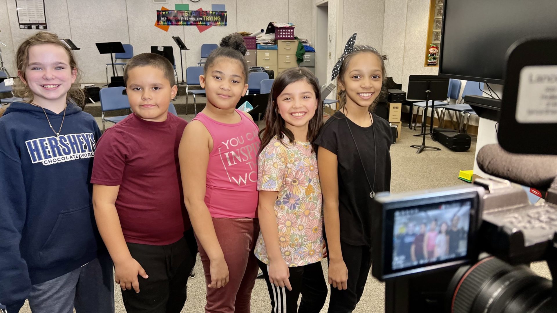a racially diverse groupd of 5 elementary students, left to right girl, boy, girl, girl, girl, and a video camera in the bottom right of the frame