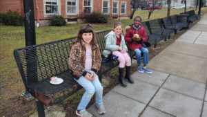 three elementary school girls - two white one black - seated on benches behind the school and eating breakfast