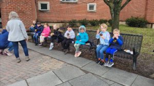 eight elementary students seated on benches behind the school and eating breakfast