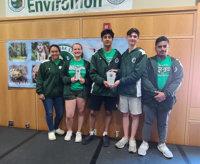 two high school girls and three high school boys wear dark green jackets and stand under an Envirothon banner