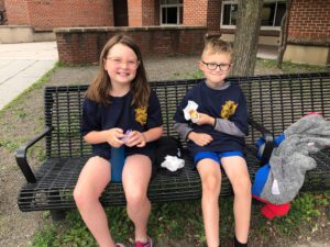 two elementary school students eating breakfast while seated on a bench outside the school