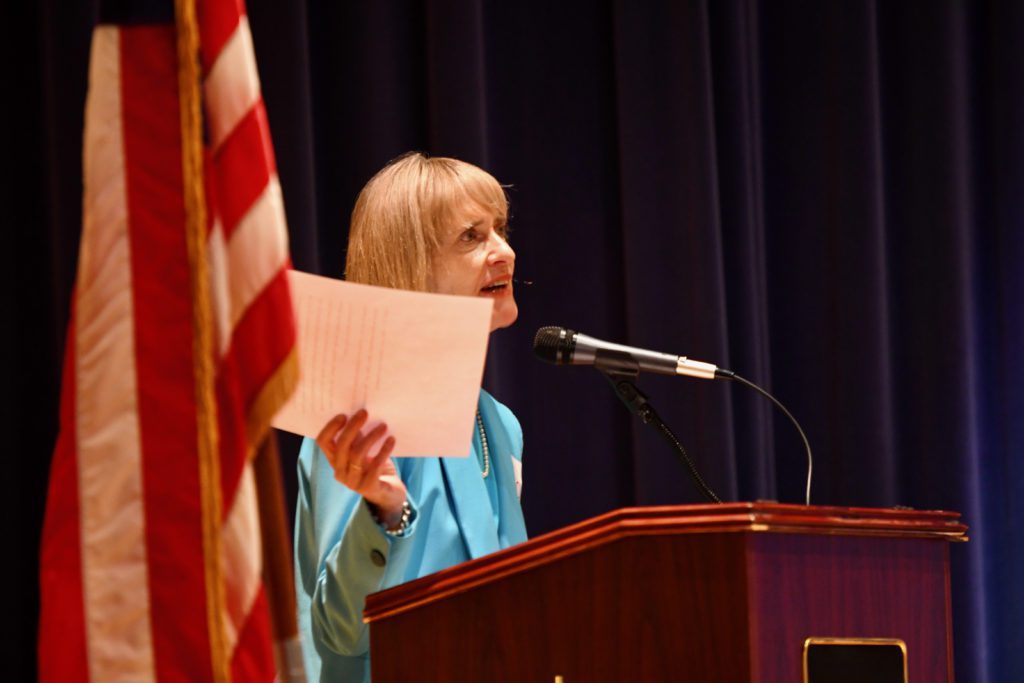 Dr. Carlee Drummer speaks at a podium in front of blue stage curtains and an American Flag