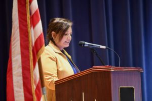 Dr. Lisamarie Spindler speaks at a podium in fron of blue stage curtains and an American Flag